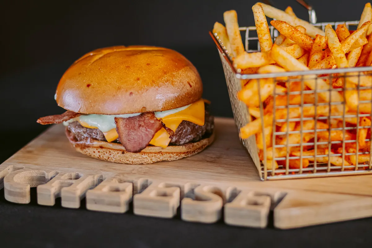 Hambúrguer com bacon e cheddar sobre tábua de madeira, ao lado de cesto de batatas fritas temperadas